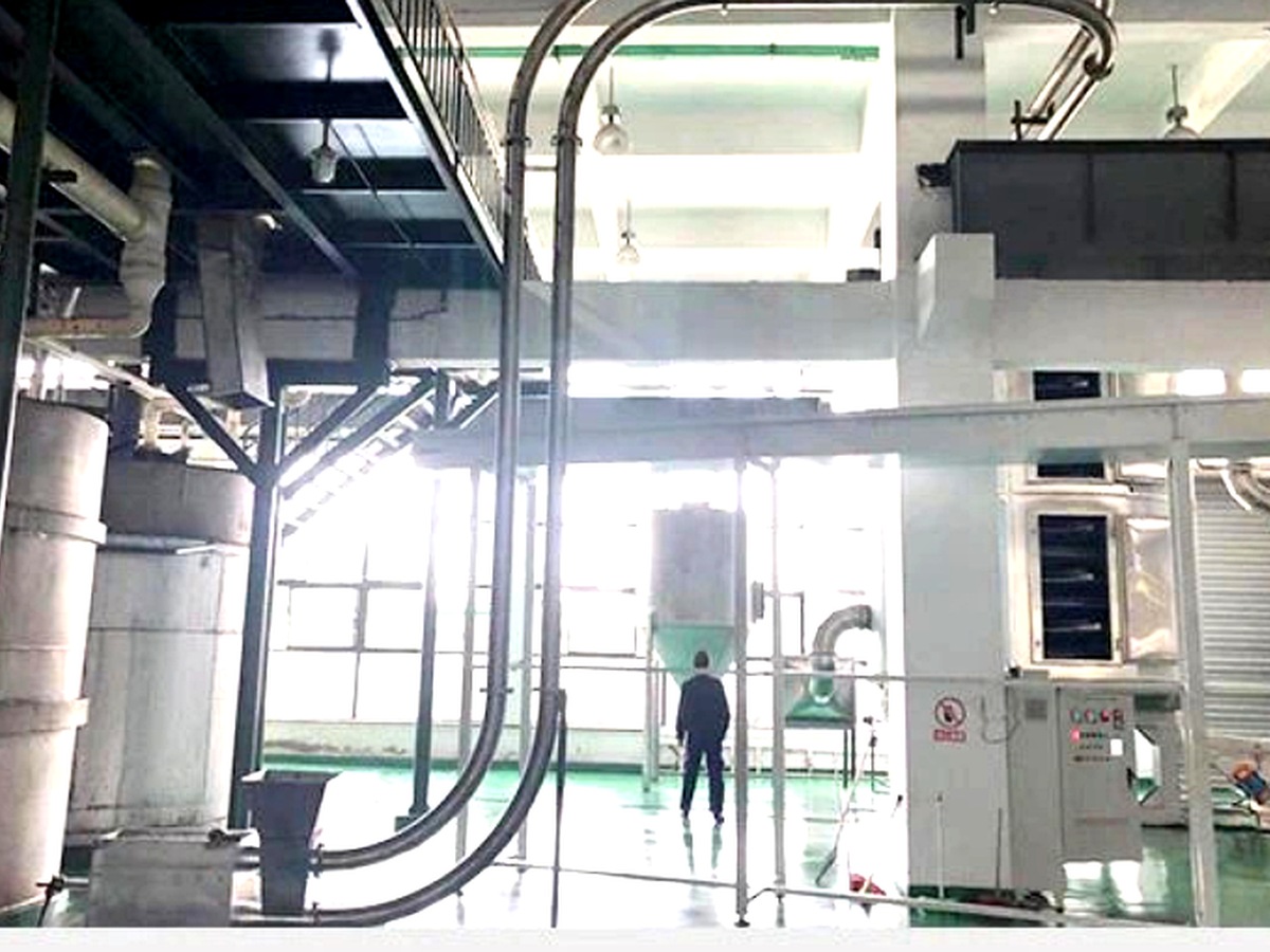Tubular drag chain conveyor installed in a large industrial warehouse showing multi-directional routing: horizontal, vertical, and curved pipe sections with blue motor drive head